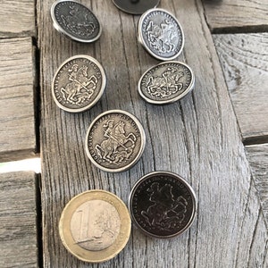 May include: A collection of antique silver-colored buttons with a raised design of a knight on horseback, along with a Euro coin. The buttons are scattered on a weathered wooden surface, showcasing their intricate details and vintage appeal.