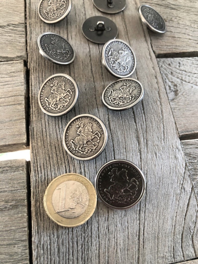May include: A collection of antique silver-colored buttons with a raised design of a knight on horseback, along with a Euro coin. The buttons are scattered on a weathered wooden surface, showcasing their intricate details and vintage appeal.
