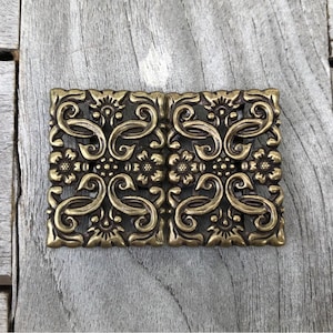 Puede incluir: Una hebilla de cinturón de metal dorado con un diseño decorativo de espirales intrincados y patrones florales.