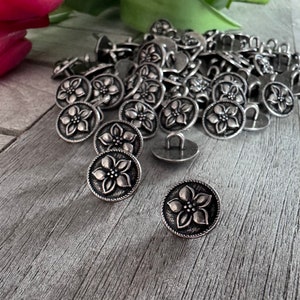 Pode incluir: Um monte de botões de metal prateado com um design floral. Os botões são redondos e têm uma flor em relevo no centro.