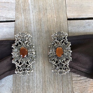 Puede incluir: Dos hebillas de cintur&oacute;n de plata adornadas con piedras de vidrio de color &aacute;mbar en el centro. Las hebillas tienen un dise&ntilde;o floral.
