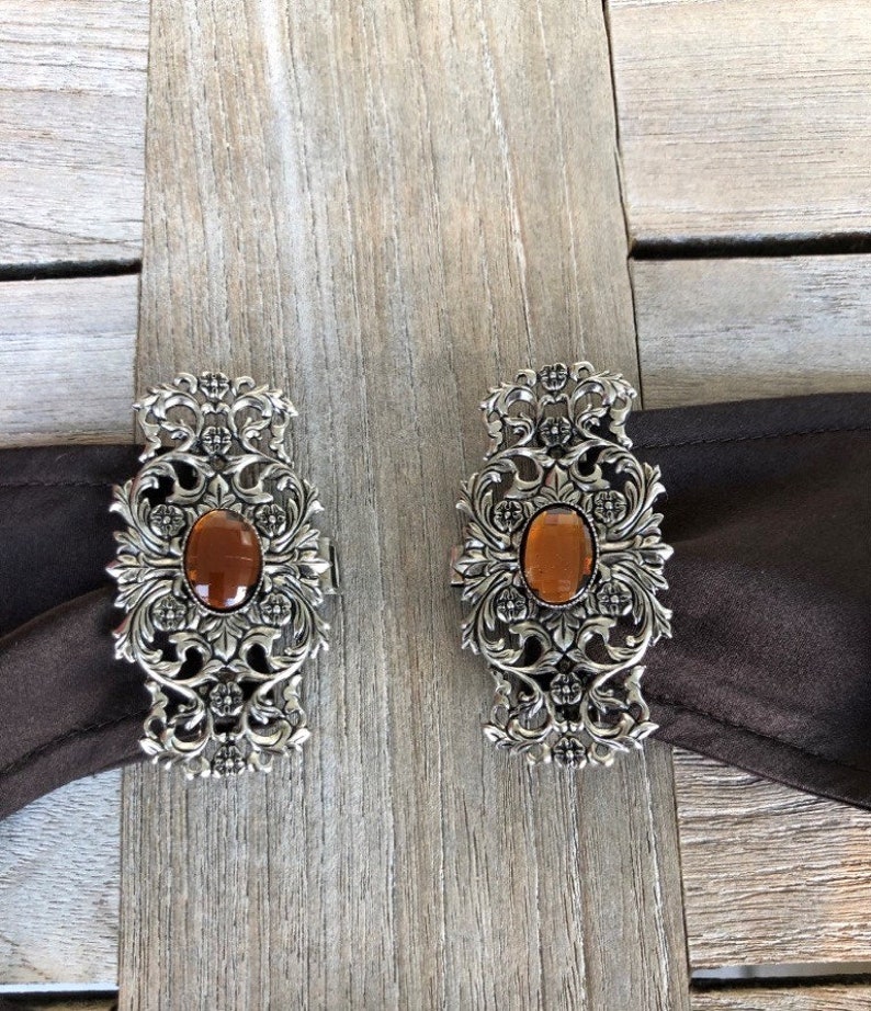 Puede incluir: Dos hebillas de cintur&oacute;n de plata adornadas con piedras de vidrio de color &aacute;mbar en el centro. Las hebillas tienen un dise&ntilde;o floral.