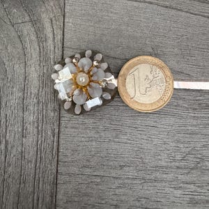 May include: A floral-shaped embellishment with a central pearl and surrounding crystals and light gray petals. A one Euro coin is next to the embellishment. The embellishment is on a gray wooden surface.