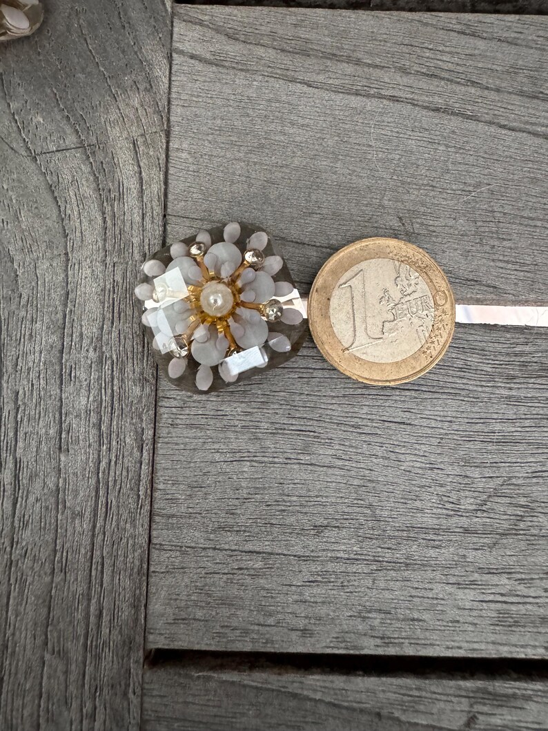 May include: A floral-shaped embellishment with a central pearl and surrounding crystals and light gray petals. A one Euro coin is next to the embellishment. The embellishment is on a gray wooden surface.
