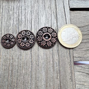 Puede incluir: Tres botones antiguos de color cobre con diseños florales y una moneda de un euro para comparar el tamaño. Los botones varían en tamaño, con un agujero central para coser. Los botones están sobre una superficie de madera envejecida.