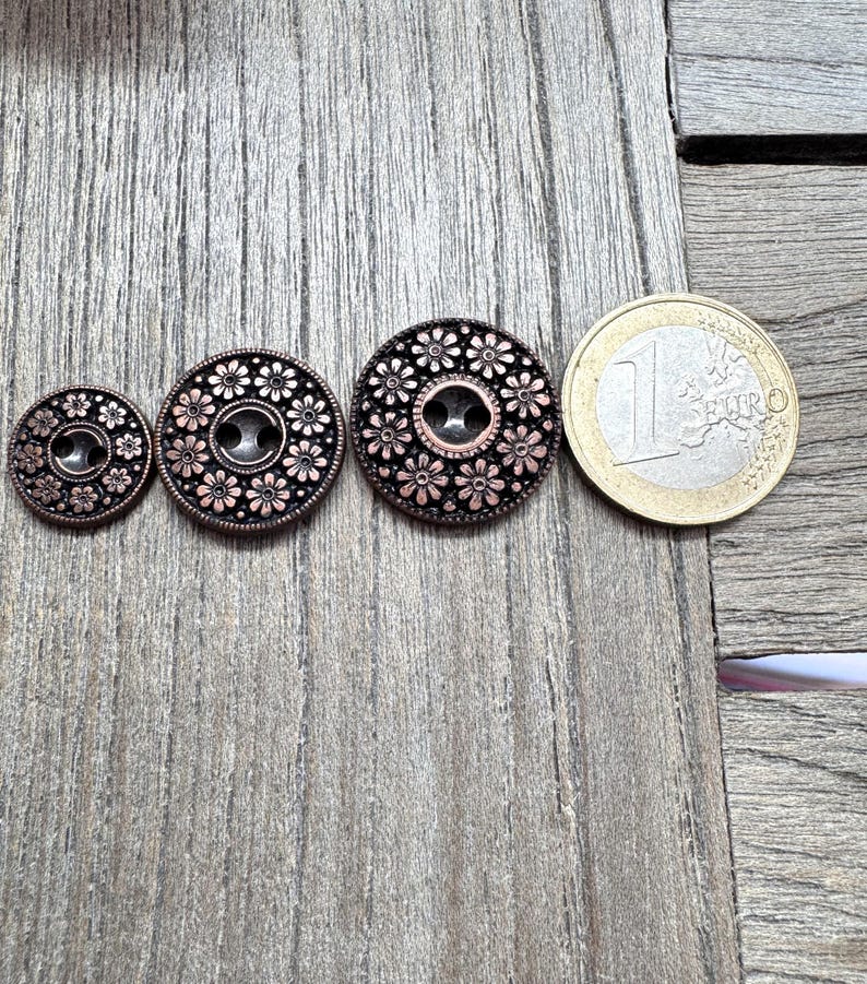 May include: Three antique copper-colored buttons with floral designs and a one Euro coin for size comparison. The buttons vary in size, with a central hole for sewing. The buttons are set against a weathered wooden surface.