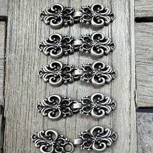 Könnte beinhalten: Sechs verzierte, silberfarbene Verschlüsse mit einem dekorativen Scroll-Design sind vertikal auf einer Holzoberfläche angeordnet. Jeder Verschluss besteht aus zwei ineinandergreifenden Teilen, ideal für Kleidung oder Accessoires.