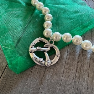 Op de afbeelding: Een witte parelketting met een zilveren pretzel-bedel. De ketting ligt op een groene stoffen achtergrond.