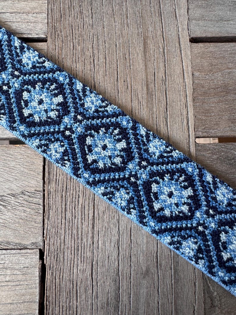 May include: A blue and white patterned textile with a geometric design. The pattern features squares and diamond shapes in shades of blue and white. The textile is placed on a wooden surface.