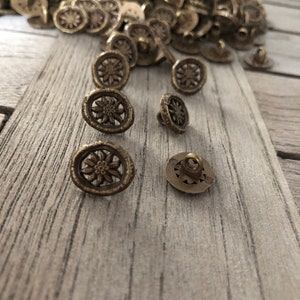 May include: A pile of antique gold-toned metal buttons with a flower design. The buttons have a shank back and are scattered on a wooden surface.