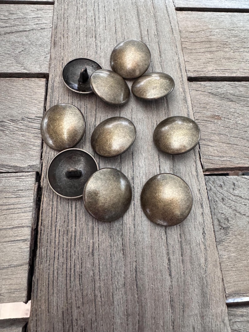 Peut inclure: Un groupe de dix boutons ronds en m&eacute;tal de couleur or antique avec un centre sur&eacute;lev&eacute;. Les boutons ont une surface lisse l&eacute;g&egrave;rement textur&eacute;e et sont dispos&eacute;s en cercle l&acirc;che sur une surface en bois.
