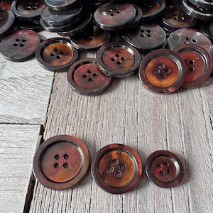 May include: A collection of brown and black buttons with four holes each. The buttons vary in size and have a marbled appearance. The buttons are arranged on a wooden surface.