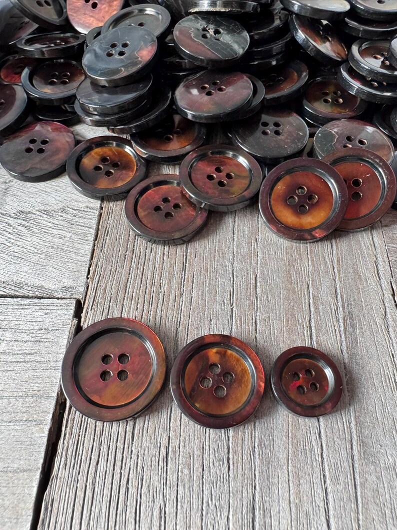 May include: A collection of brown and black buttons with four holes each. The buttons vary in size and have a marbled appearance. The buttons are arranged on a wooden surface.
