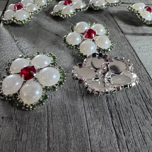 Peut inclure: Un ensemble de boutons en forme de fleur de perle blanche avec des centres en strass rouges et des accents en strass verts. Les boutons sont disposés sur une surface en bois.