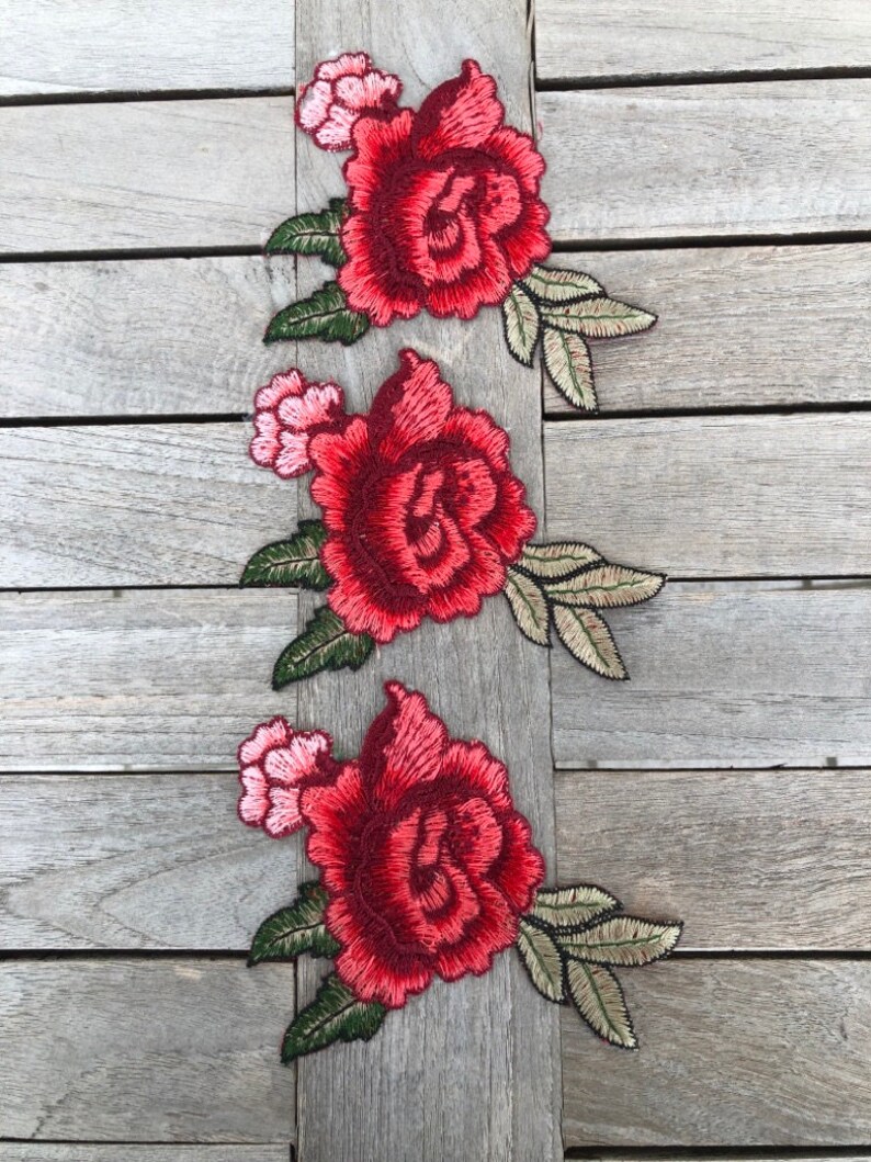 May include: Three red embroidered rose appliques with green leaves on a wood background.