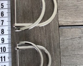 Puede incluir: Dos hebillas de metal plateado en forma de D, cada una con un diseño curvo, se muestran sobre una superficie de madera. Las hebillas están colocadas verticalmente, con una cinta métrica al lado. Medidas en centímetros.