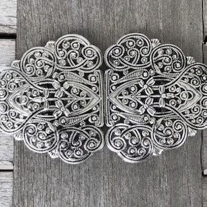 Puede incluir: Una hebilla de cinturón de filigrana plateada con un diseño decorativo. La hebilla está hecha de metal y tiene un patrón detallado.