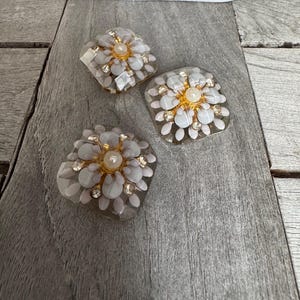 May include: Three decorative floral-shaped embellishments. Each features a central pearl surrounded by gold-toned accents and clear, petal-like elements. The embellishments are set with small, clear crystals and are displayed on a weathered, gray wooden surface.