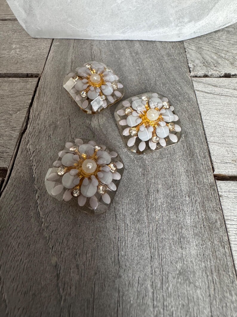 May include: Three decorative floral-shaped embellishments. Each features a central pearl surrounded by gold-toned accents and clear, petal-like elements. The embellishments are set with small, clear crystals and are displayed on a weathered, gray wooden surface.
