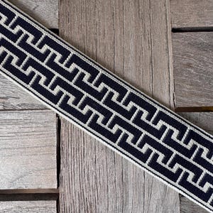 Puede incluir: Cinta estampada en azul marino y blanco con un diseño geométrico. La cinta está colocada en diagonal sobre una superficie de madera. El patrón es una serie repetida de formas de llave griega estilizadas y conectadas.