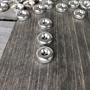 Peut inclure: Gros plan sur trois boutons en métal argenté à quatre trous chacun, disposés en rangée verticale sur une surface en bois foncé. Les boutons sont brillants et ont une finition lisse.