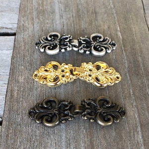 May include: Three ornate metal clasps in silver, gold, and bronze. Each clasp has a decorative, intricate design with a loop on each side.