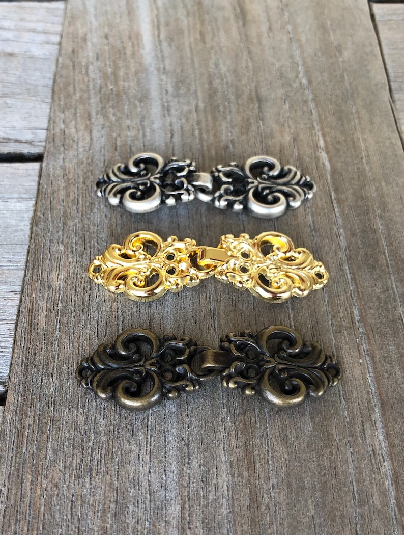 May include: Three ornate metal clasps in silver, gold, and bronze. Each clasp has a decorative, intricate design with a loop on each side.