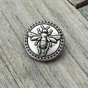 May include: A silver-colored button with a detailed bee design in the center. The bee is surrounded by a chain-like border. The button is round and appears to be made of metal. The background is a wooden surface.