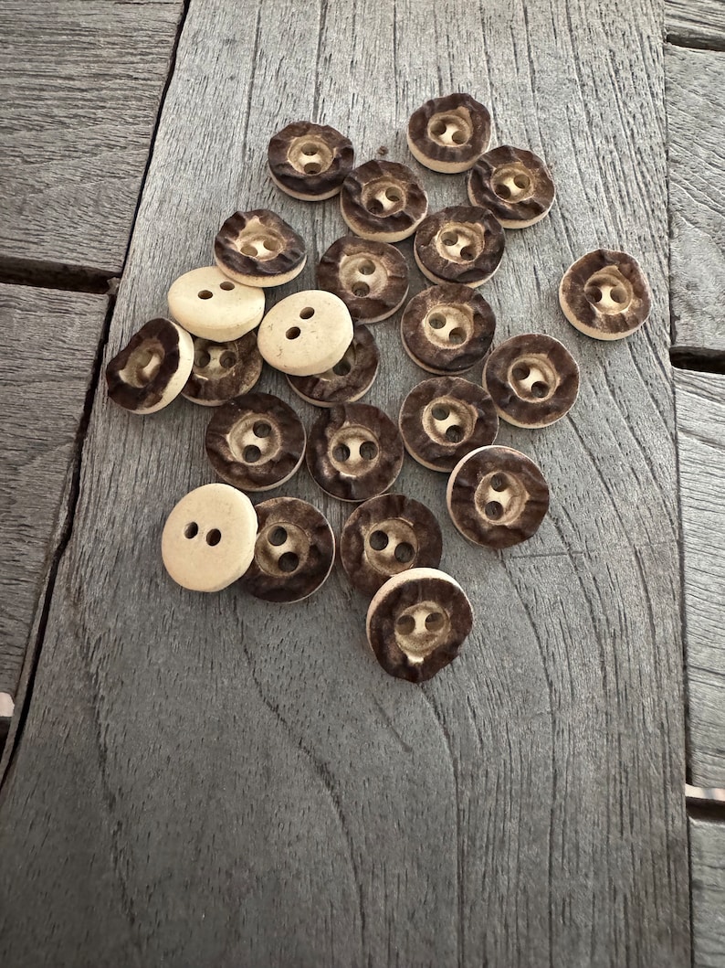 Peut inclure: Un ensemble de boutons en bois brun et beige avec deux trous chacun. Les boutons sont dispos&eacute;s en pile sur une surface en bois.