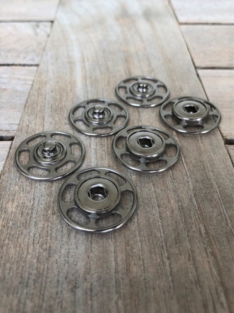 May include: Six silver metal snap fasteners with a circular design. The fasteners have a raised center and a series of small holes around the edge.