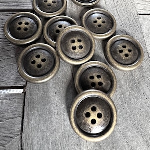 Könnte beinhalten: Zehn antike Metallknöpfe mit jeweils vier Löchern. Die Knöpfe sind kreuzförmig auf einer Holzoberfläche angeordnet. Die Knöpfe sind dunkelgolden mit einer leicht abgenutzten Oberfläche.