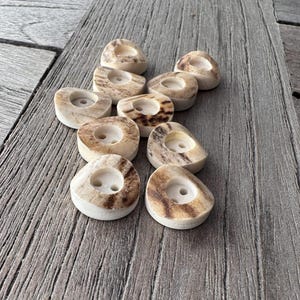 May include: A collection of small, off-white candle holders with a natural, speckled brown pattern. Each holder has a circular indentation for a candle. The holders are arranged on a weathered wooden surface.