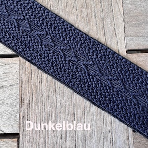 Puede incluir: Cinta tejida azul marino con un patrón texturizado y el texto "Dunkelblau". La cinta está en ángulo sobre una superficie de madera. El diseño de la cinta presenta un patrón geométrico repetitivo.