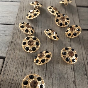 Op de afbeelding: Een set van 12 goudkleurige knopen met een zwart en bruin strass-ontwerp. Elke knop is rond met een verhoogd midden en een decoratieve rand.