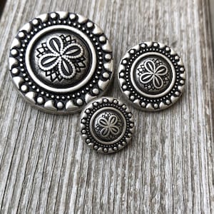 Peut inclure: Trois boutons en métal argenté avec un motif floral décoratif. Les boutons sont de tailles différentes et ont un bord surélevé et texturé.