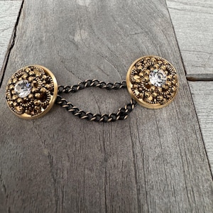 May include: A pair of gold-toned metal buttons with clear rhinestone centres connected by a black chain. The buttons have a decorative, beaded design.