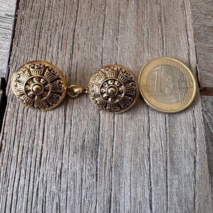 May include: Gold-coloured metal cufflinks with a detailed circular design. The cufflinks are linked by a small chain and are shown next to a one Euro coin for size comparison. The design features a central circle surrounded by intricate patterns.