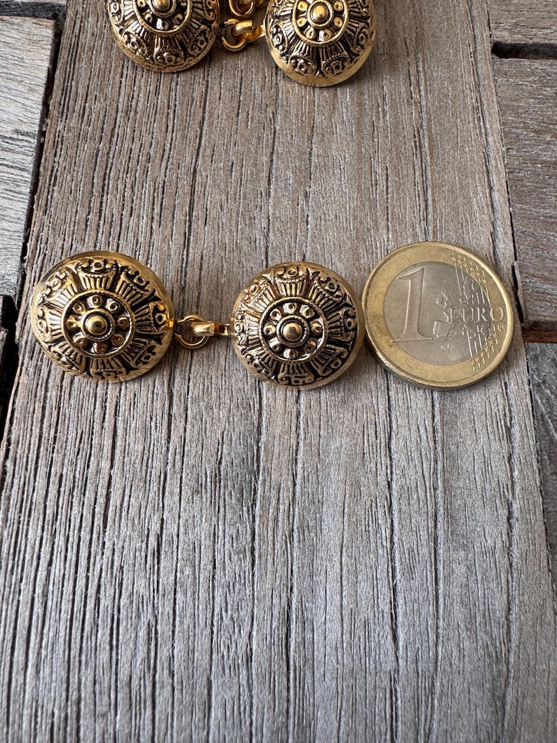 May include: Gold-coloured metal cufflinks with a detailed circular design. The cufflinks are linked by a small chain and are shown next to a one Euro coin for size comparison. The design features a central circle surrounded by intricate patterns.