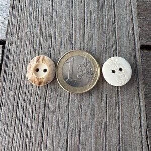 May include: Two buttons and a Euro coin are displayed on a weathered wooden surface. One button is light brown with two holes, the other is off-white with two holes. The coin is gold and silver, with the number "1" visible.