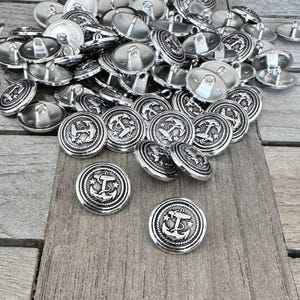 Op de afbeelding: Een stapel zilveren metalen knopen met een ankerontwerp. De knopen zijn rond en hebben een verhoogde rand.