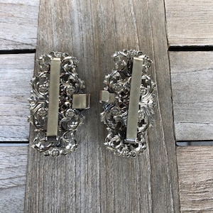 Puede incluir: Dos hebillas de cintur&oacute;n de metal plateadas con dise&ntilde;os florales ornamentados. Las hebillas tienen una forma rectangular y un acabado liso y pulido.