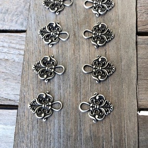 以下が含まれることがあります： 木製の表面に、花柄の装飾が施された銀色の金属製の留め具が8個並んでいます。留め具の上部にはループが付いており、4個ずつ2列に並んでいます。