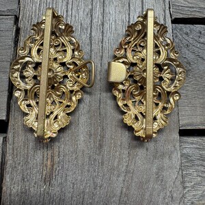 May include: Two ornate, antique-style brass belt buckles. Each buckle features a detailed, symmetrical floral and scroll design. The buckles are set against a weathered, gray wooden surface, highlighting their intricate details and aged appearance.
