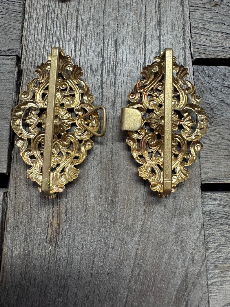 May include: Two ornate, antique-style brass belt buckles. Each buckle features a detailed, symmetrical floral and scroll design. The buckles are set against a weathered, gray wooden surface, highlighting their intricate details and aged appearance.