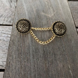 Puede incluir: Broche de botón dorado de estilo vintage con dos botones redondos y texturizados unidos por una delicada cadena. Los botones tienen una superficie detallada y estampada. El broche se encuentra sobre un fondo de madera rústica y envejecida.