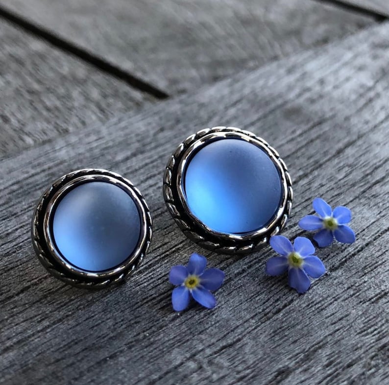 May include: A pair of silver stud earrings with a light blue glass cabochon in each. The earrings are on a wooden surface with small blue flowers.