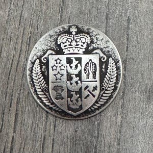Puede incluir: Una moneda plateada con un escudo de armas que presenta una corona, un escudo dividido en cuatro secciones y un diseño de hoja de helecho.