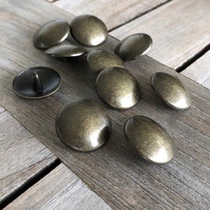 Peut inclure: Un groupe de dix boutons en m&eacute;tal de couleur or antique. Les boutons sont ronds et l&eacute;g&egrave;rement bomb&eacute;s. Ils sont dispos&eacute;s sur une surface en bois.