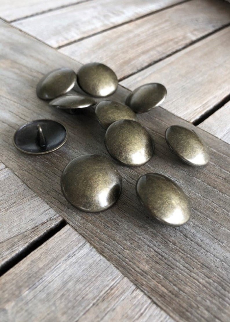 Peut inclure: Un groupe de dix boutons en m&eacute;tal de couleur or antique. Les boutons sont ronds et l&eacute;g&egrave;rement bomb&eacute;s. Ils sont dispos&eacute;s sur une surface en bois.