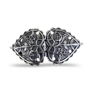 Könnte beinhalten: Eine silberne herzförmige Schnalle mit filigranen Blumendesigns. Die Schnalle ist aus Metall und hat eine polierte Oberfläche.